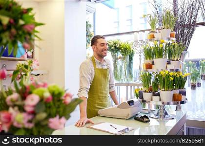 people, sale, retail, business and floristry concept - happy smiling florist man with clipboard and cashbox standing at flower shop counter