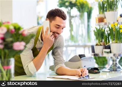 people, sale, retail, business and floristry concept - happy smiling florist man calling on smartphone and making notes to clipboard at flower shop counter. florist man calling on smartphone at flower shop