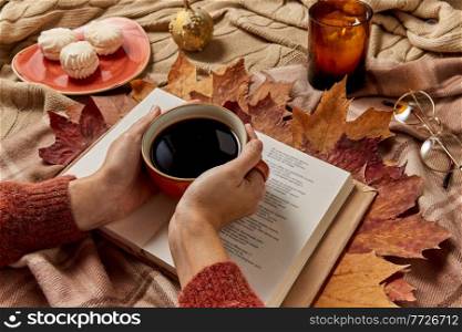 people, season and leisure concept - close up of hands holding cup of coffee, book, autumn leaves, meringues and candle burning on warm blankets at home. hands with cup of coffee, autumn leaves and book