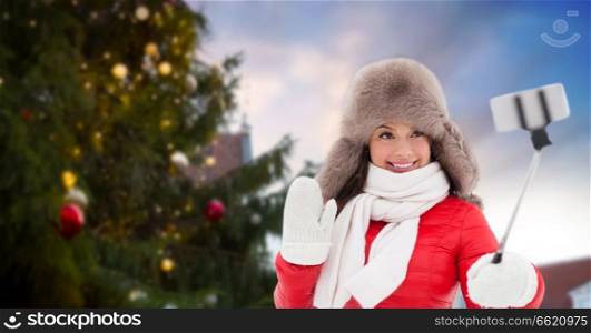 people, season and leisure concept - happy smiling woman in winter fur hat taking selfie by smartphone over christmas tree at tallinn old town hall square background. woman taking selfie over christmas tree