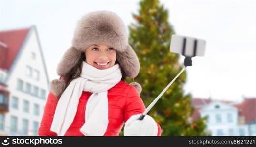 people, season and leisure concept - happy smiling woman in winter fur hat taking selfie by smartphone over christmas tree at tallinn old town hall square background. woman taking selfie over christmas tree