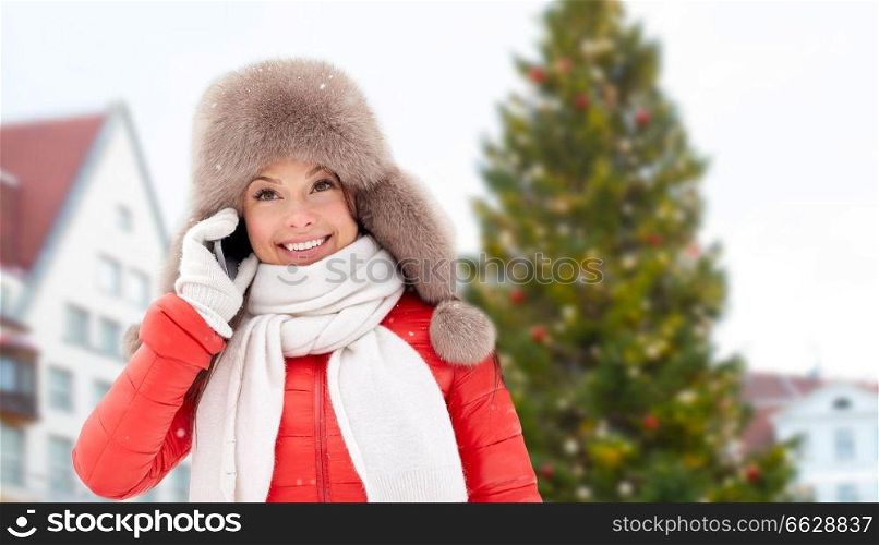 people, season and leisure concept - happy smiling woman in winter fur hat calling on smartphone over christmas tree at tallinn old town hall square background. woman calling on smartphone over christmas tree