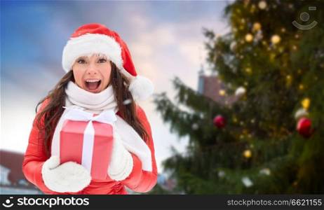 people, season and leisure concept - happy woman in santa hat with gift over christmas tree at tallinn old town hall square background. happy woman with gift over christmas tree