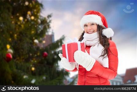 people, season and leisure concept - happy woman in santa hat with gift over christmas tree at tallinn old town hall square background. happy woman with gift over christmas tree