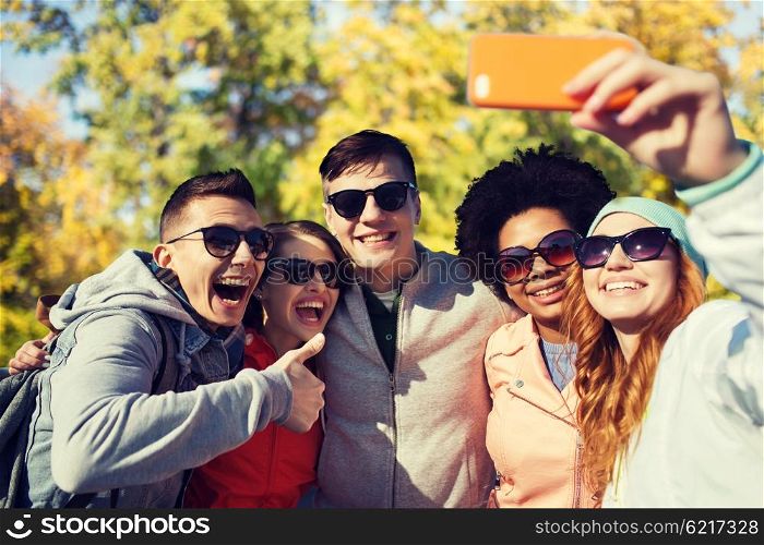 people, season, friendship and technology concept - group of smiling teenage friends taking selfie with smartphone and showing thumbs up over autumn park background