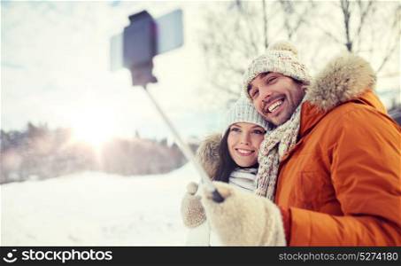 people, season, love, technology and leisure concept - happy couple taking picture with smartphone selfie stick on over winter background. happy couple taking selfie by smartphone in winter