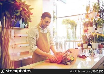 people, shopping, sale, floristry and consumerism concept - happy smiling florist man wrapping flowers in paper at flower shop. florist wrapping flowers in paper at flower shop