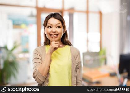 people, silence and corporate secret concept - happy asian young woman with finger on lips over office background. happy asian woman with finger on lips