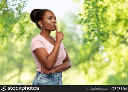 people, silence and secret concept - african american young woman with finger on lips over green natural background. african american woman with finger on lips