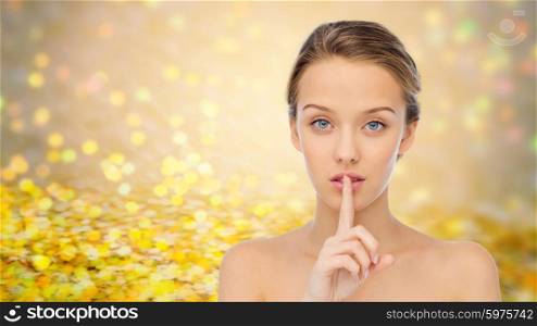 people, silence, secret, gesture and beauty concept - beautiful young woman holding finger on lips over golden glitter or holidays lights background