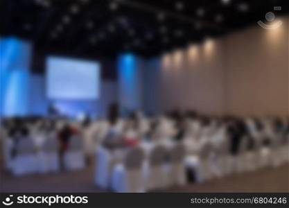 people sit on rows of chairs in large conference convention hall during meeting seminar, defocused blur background