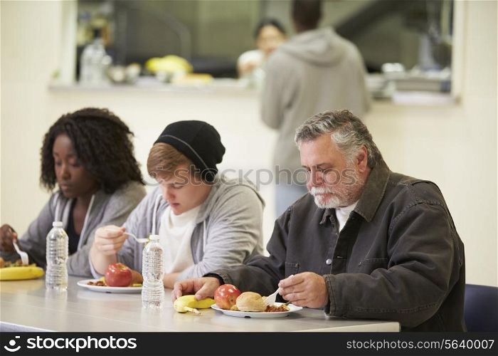 People Sitting At Table Eating Food In Homeless Shelter