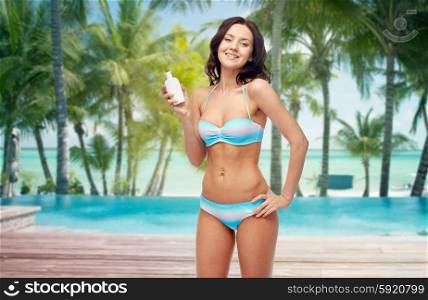 people, skincare, tanning, summer and travel concept - happy young woman in bikini swimsuit holding sunscreen bottle over tropical beach with palm trees and pool at hotel resort background