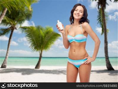 people, skincare, tanning, summer and travel concept - happy young woman in bikini swimsuit holding sunscreen bottle over tropical beach with palm trees background