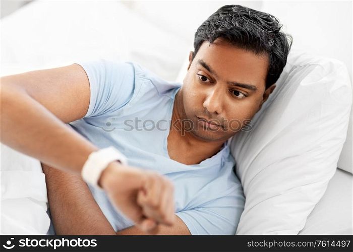 people, sleeping and rest concept - indian man looking at smart watch in bed at home. indian man looking at smart watch in bed at home