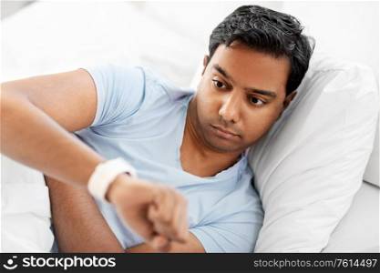 people, sleeping and rest concept - indian man looking at smart watch in bed at home. indian man looking at smart watch in bed at home