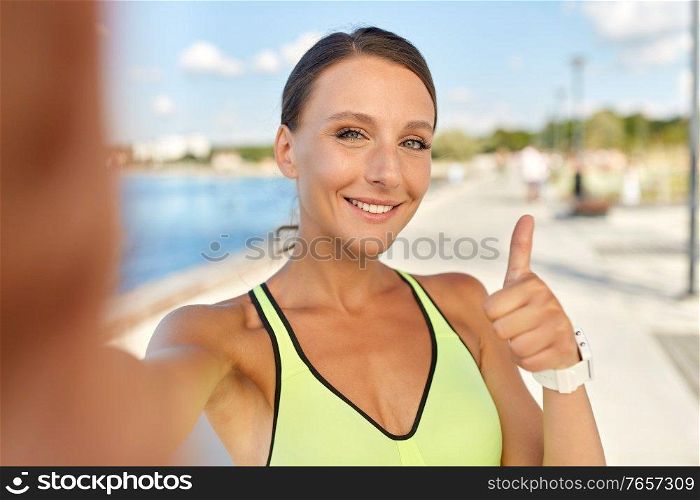 people, sport and fitness concept - portrait of smiling young sporty woman taking selfie picture and showing thumbs up outdoors on sea promenade. sporty woman taking selfie and showing thumbs up
