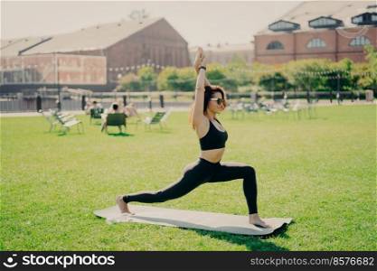 People sport and recreation concept. Sporty woman has healthy body raises arms exercises on karemat outdoors wears cropped top and leggings enjoys workout outside. Sportswoman in good shape.