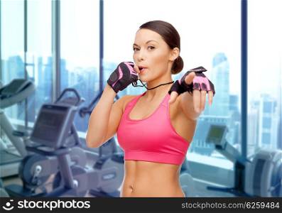 people, sport, fitness and healthy lifestyle concept - asian woman coach blowing whistle over gym machines background