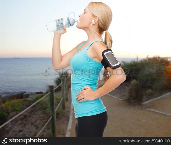 people, sport, fitness, jogging and technology concept - happy woman with smartphone and earphones listening to music and drinking water from bottle over beach sunset background