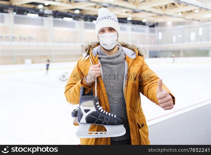 people, sport, gesture and leisure concept - young man wearing face protective medical mask for protection from virus disease with ice-skates showing thumbs up on skating rink. man in mask showing thumbs up on skating rink