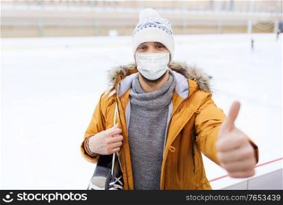 people, sport, gesture and leisure concept - young man wearing face protective medical mask for protection from virus disease with ice-skates showing thumbs up on skating rink. man in mask showing thumbs up on skating rink