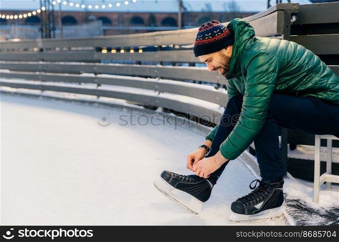 People, sport, winter, leisure concept. Smiling delightful male laces up skates, going to practice on ice ring, being in good mood, likes winter and go skating with best friends. Hobby concept