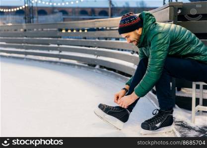 People, sport, winter, leisure concept. Smiling delightful male laces up skates, going to practice on ice ring, being in good mood, likes winter and go skating with best friends. Hobby concept