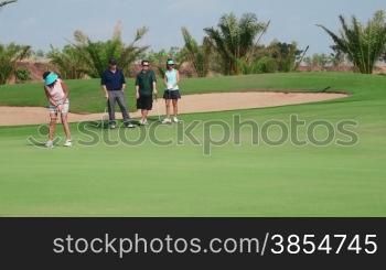 People, sports, leisure activities, recreation and lifestyle, golf in country club during summer holidays. Asian woman hitting ball with iron in golf course near hole, friends watching. 15of30