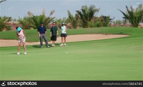 People, sports, leisure activities, recreation and lifestyle, golf in country club during summer holidays. Asian woman hitting ball with iron in golf course near hole, friends watching. 15of30