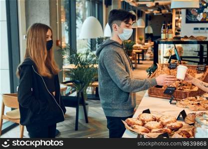 People standing in a queue to buy coffee and pastry in the coffee shop to go. Man taking coffee cup from barista. People standing at counter wearing the face masks to avoid virus infection and to prevent the spread of disease in time of coronavirus