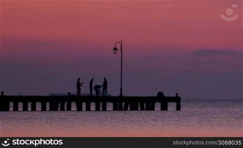People standing silhouetted against a pink sky on a pier at sunset viewed across a calm sea