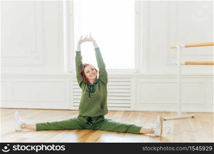 People, strength, fitness, gymnastics concept. Glad sportswoman with cheerful face, raises hands, spreads legs and does splits, stretches in studio. Young dancer has work out, dressed in tracksuit