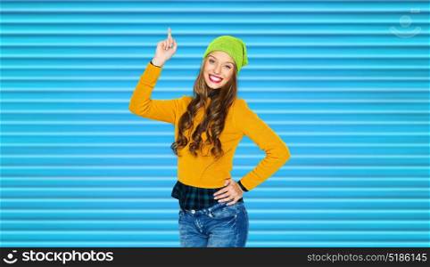 people, style and fashion concept - happy young woman or teen girl in casual clothes and hipster hat pointing finger up over blue ribbed background. happy young woman or teen girl pointing finger up