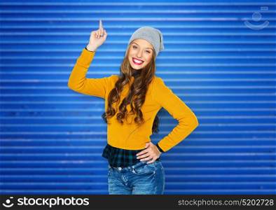 people, style and fashion concept - happy young woman or teen girl in casual clothes and hipster hat pointing finger up over blue ribbed background. happy young woman or teen girl pointing finger up