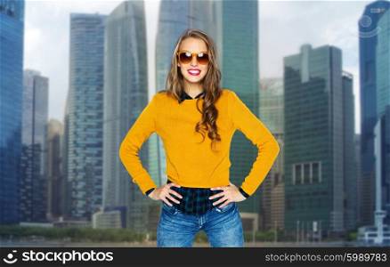 people, style and fashion concept - happy young woman or teen girl in casual clothes and sunglasses over city buildings and skyscrapers background