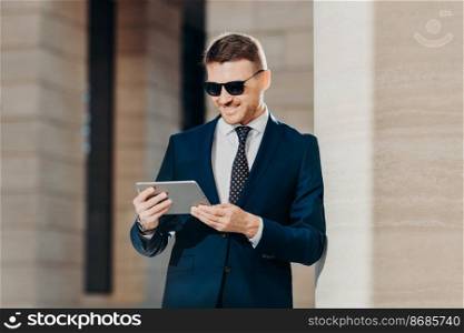 People, style, business and technology concept. Horizontal shot of attractive young businessman reads important information on tablet computer, connected to wifi, wears trendy shades and suit