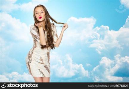 people, style, holidays, hairstyle and fashion concept - happy young woman or teen girl in fancy dress with sequins and long wavy hair sending blow kiss over blue sky and clouds background