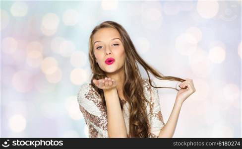 people, style, holidays, hairstyle and fashion concept - happy young woman or teen girl in fancy dress with sequins and long wavy hair sending blow kiss over holidays lights background