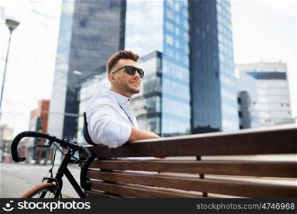 people, style, leisure and lifestyle - happy smiling young man with bicycle sitting on city bench