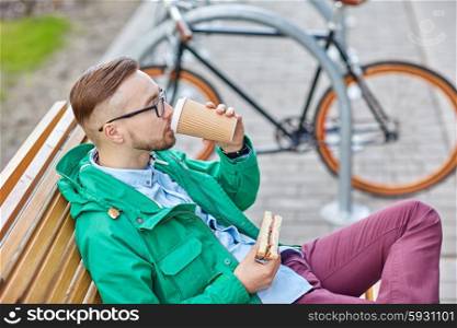 people, style, leisure and lifestyle - happy young hipster man drinking coffee cup and eating sandwich on city street