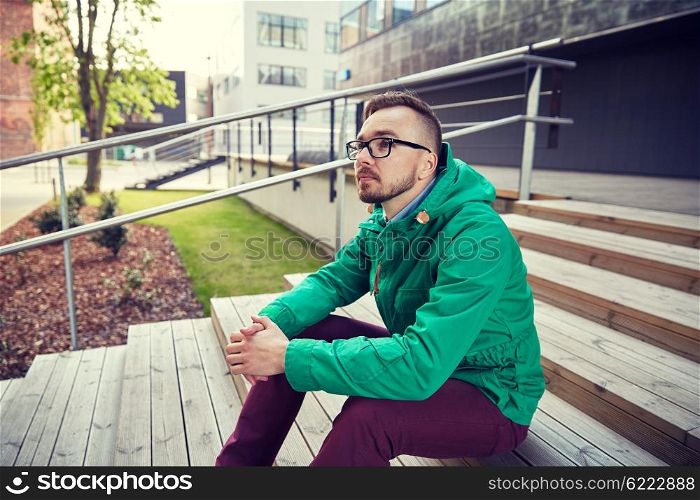 people, style, leisure and lifestyle - happy young hipster man in eyeglasses and jacket sitting on stairs in city