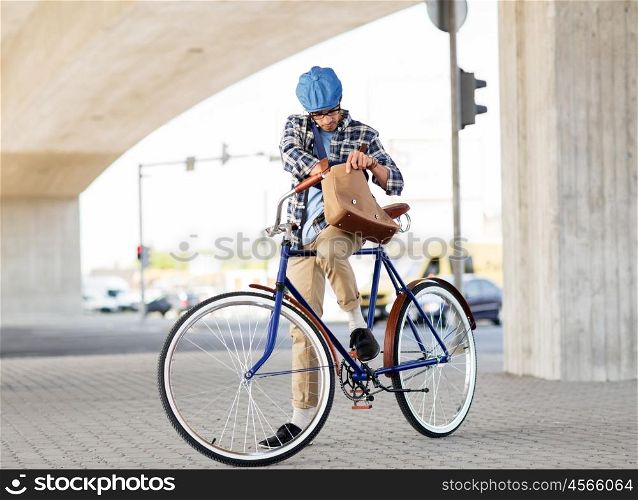 people, style, leisure and lifestyle - hipster man lookir for something in his shoulder bag riding fixed gear bike on city street