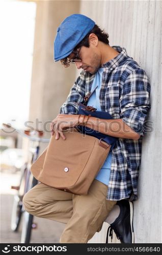 people, style, leisure and lifestyle - hipster man lookir for something in his shoulder bag and fixed gear bike on city street