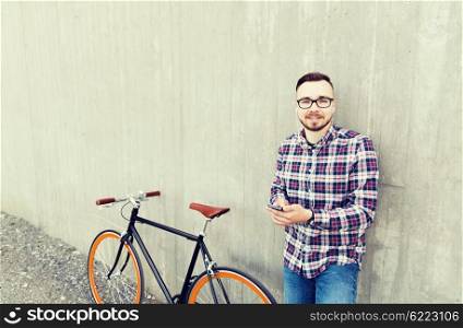 people, style, technology, leisure and lifestyle - happy young hipster man in earphones with smartphone and fixed gear bike listening to music on city street