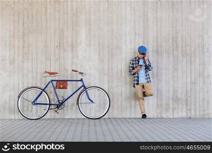 people, style, technology, leisure and lifestyle - young hipster man in earphones with smartphone and fixed gear bike listening to music at city street wall