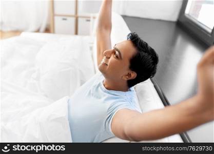 people, success and rest concept - happy smiling indian man lying in bed at home. happy smiling indian man lying in bed at home