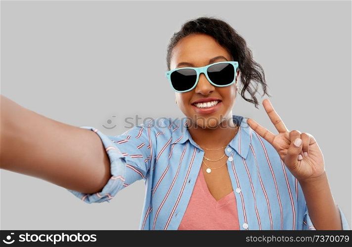 people, summer and gesture concept - happy african american young woman in sunglasses taking selfie and showing peace hand sign over grey background. african american woman in sunglasses taking selfie