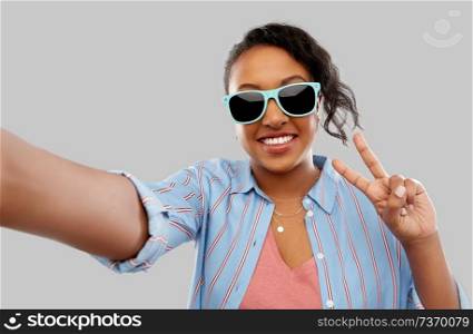 people, summer and gesture concept - happy african american young woman in sunglasses taking selfie and showing peace hand sign over grey background. african american woman in sunglasses taking selfie