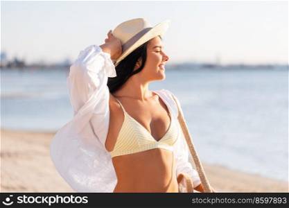 people, summer and leisure concept - portrait of young woman in bikini swimsuit, white shirt and straw hat with bag on beach. portrait of woman in bikini and hat on beach